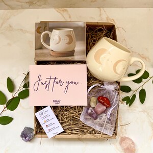 May include: A gift box containing a cream-coloured mug with moon and star designs, a pink card with "Just for you..." text, and a small bag of crystals. The box also contains a mug on a cork coaster. The theme is self-care.