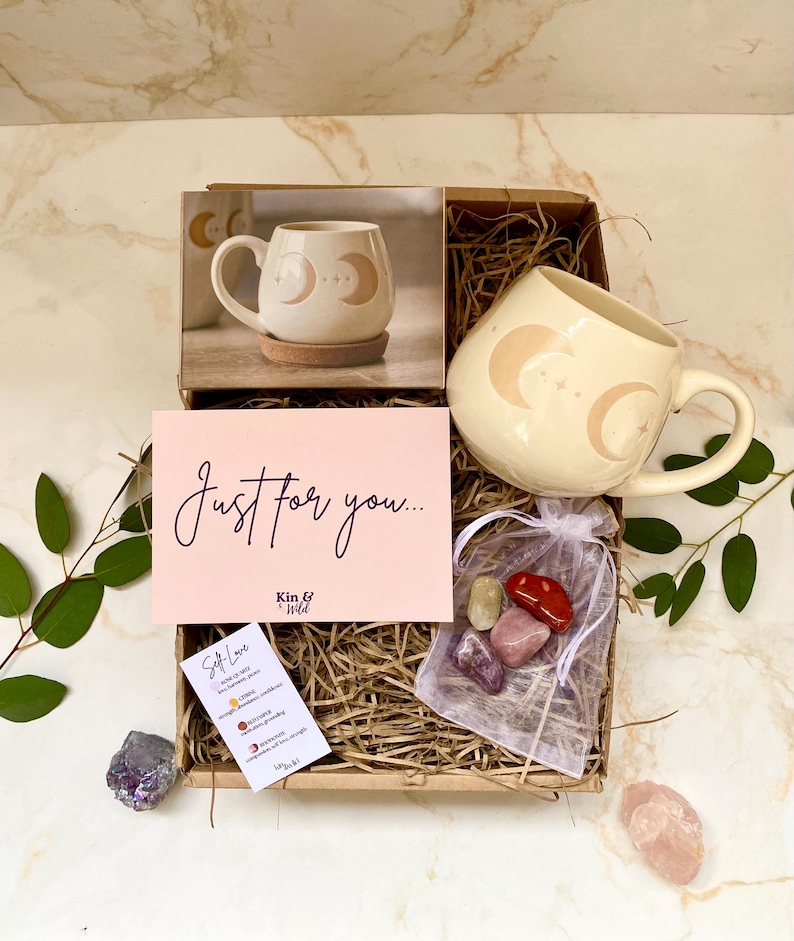 May include: A gift box containing a cream-coloured mug with moon and star designs, a pink card with "Just for you..." text, and a small bag of crystals. The box also contains a mug on a cork coaster. The theme is self-care.
