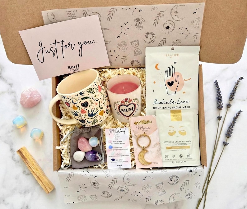 May include: A gift box filled with self-care items. Includes a floral mug, a candle with "MUM" on it, a face mask, eye masks, crystals, a moon-shaped keychain, and a card that says "Just for you...". The box is lined with patterned paper.