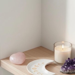 Peut inclure: Un cristal rose en forme de c&oelig;ur, un plat en c&eacute;ramique blanc en forme de croissant de lune avec des motifs de phases lunaires dor&eacute;es, une bougie allum&eacute;e dans un verre et un amas de cristaux d'am&eacute;thyste violets sont dispos&eacute;s sur une &eacute;tag&egrave;re en bois.