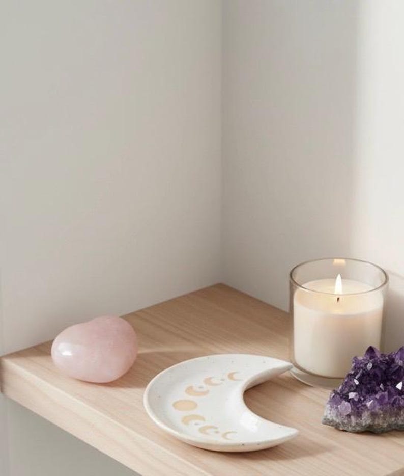 Peut inclure: Un cristal rose en forme de c&oelig;ur, un plat en c&eacute;ramique blanc en forme de croissant de lune avec des motifs de phases lunaires dor&eacute;es, une bougie allum&eacute;e dans un verre et un amas de cristaux d'am&eacute;thyste violets sont dispos&eacute;s sur une &eacute;tag&egrave;re en bois.