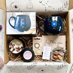 Op de afbeelding: Een open cadeauverpakking gevuld met zelfzorgartikelen. Inclusief een blauwe mok met een oogontwerp, kristallen, een kaars, een sleutelhanger en een kaart met de tekst "Just for you...". De doos is versierd met hemelse ontwerpen.