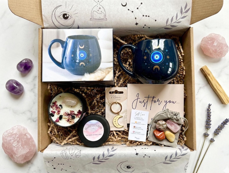 Op de afbeelding: Een open cadeauverpakking gevuld met zelfzorgartikelen. Inclusief een blauwe mok met een oogontwerp, kristallen, een kaars, een sleutelhanger en een kaart met de tekst "Just for you...". De doos is versierd met hemelse ontwerpen.