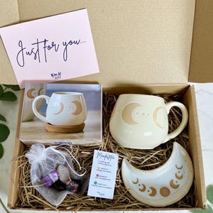 Peut inclure: Coffret cadeau contenant une tasse cr&egrave;me avec un motif de croissant de lune, un plat assorti en forme de lune, un petit sachet de cristaux et une carte avec l'inscription "Juste pour vous". Les articles sont dispos&eacute;s sur de la paille.