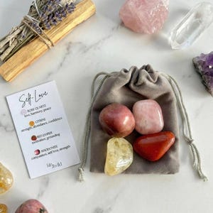 Op de afbeelding: Een verzameling kristallen en stenen, waaronder rozenkwarts, citrien en rode jaspis, samen met een kleine grijze zak met trekkoord. Er is ook een kaart met de woorden "Self Love" en kristaleigenschappen, samen met een bosje gedroogde lavendel en een palo santo-stok.