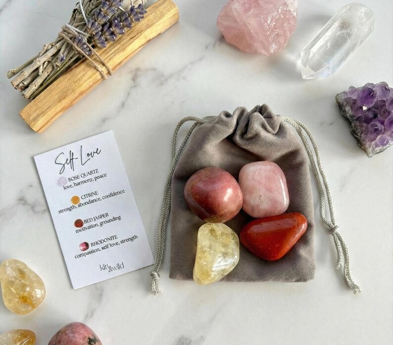 Op de afbeelding: Een verzameling kristallen en stenen, waaronder rozenkwarts, citrien en rode jaspis, samen met een kleine grijze zak met trekkoord. Er is ook een kaart met de woorden "Self Love" en kristaleigenschappen, samen met een bosje gedroogde lavendel en een palo santo-stok.
