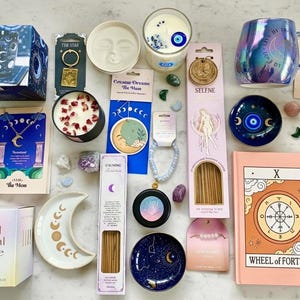 May include: A collection of celestial-themed products, including candles, mugs, incense, a necklace, a bracelet, a journal, and decorative dishes. The colour scheme features blues, pinks, and gold accents, with moon and star motifs.