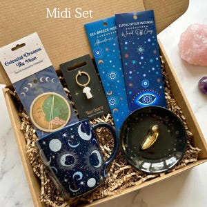May include: A gift box containing a dark blue mug with moon and star patterns, a Celestial Dreams air freshener, a rose quartz mushroom keychain, two packs of incense, a black trinket dish with a gold object, a rose quartz stone, and palo santo.