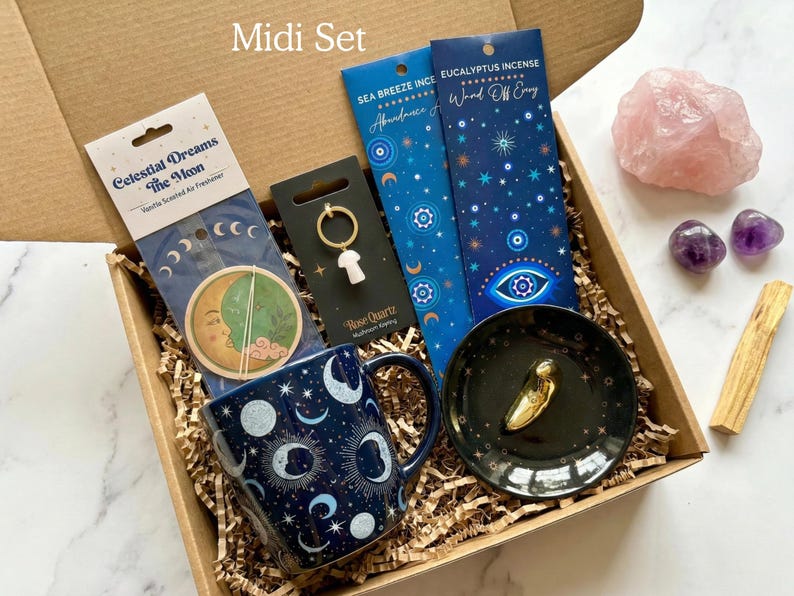 May include: A gift box containing a dark blue mug with moon and star patterns, a Celestial Dreams air freshener, a rose quartz mushroom keychain, two packs of incense, a black trinket dish with a gold object, a rose quartz stone, and palo santo.