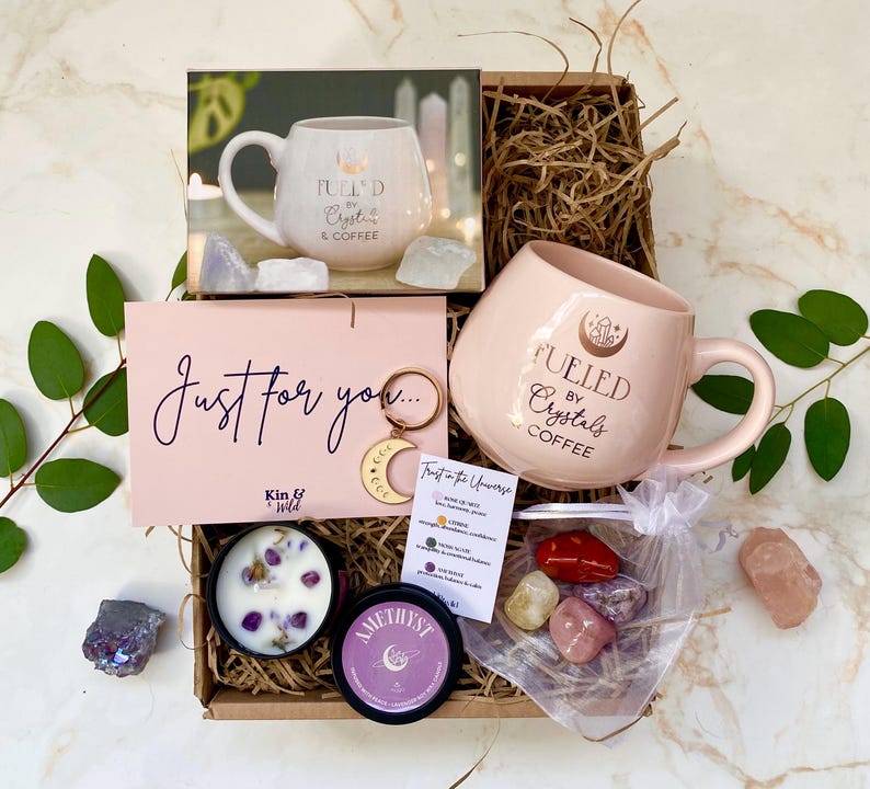 May include: A gift box containing a pink mug with the text "Fueled by Crystals & Coffee", a candle, crystals, and a card reading "Just for you". A gold moon keychain is also included.