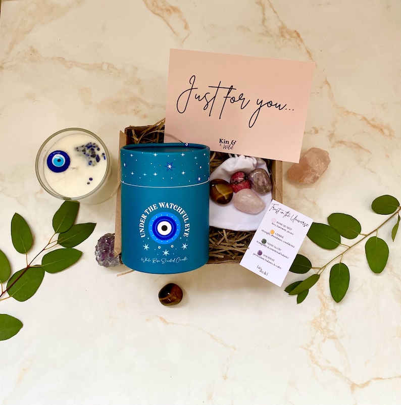 Puede incluir: Un juego de regalo con una vela azul con un dise&ntilde;o de ojo turco, una peque&ntilde;a bolsa de cristales y una tarjeta que dice "Just for you...". La vela est&aacute; en un recipiente cil&iacute;ndrico con el texto "UNDER THE WATCHFUL EYE".