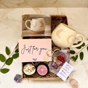 May include: A gift box with a cream-coloured mug featuring a crescent moon design, a candle, crystals, and a card reading "Just for you...". The box also contains a small bag of crystals and a small keychain.