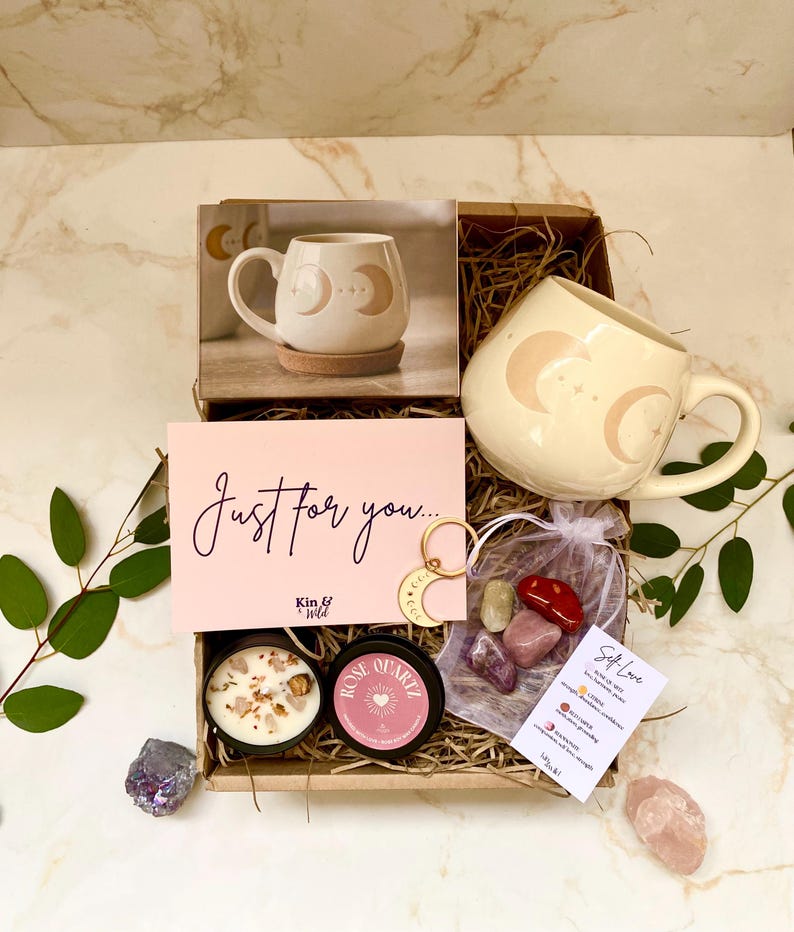 May include: A gift box with a cream-coloured mug featuring a crescent moon design, a candle, crystals, and a card reading "Just for you...". The box also contains a small bag of crystals and a small keychain.