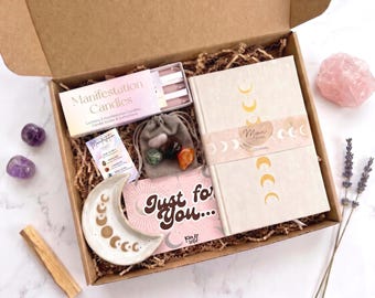 May include: A gift box containing a Moon Phase journal, a crescent moon ceramic dish, a box of Manifestation Candles, a small gray drawstring pouch with assorted crystals, a Just for You card, two amethyst stones, a rose quartz, and lavender.