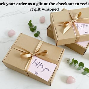 May include: Kraft gift boxes tied with gold satin ribbon and bows. Each box has a pink card with the words "Just for you..." in cursive. The boxes are arranged with pink stones and greenery on a marble surface.