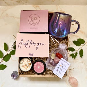 May include: A gift box containing an iridescent mug, a candle, and crystals. The mug has the text "Live by the Sun, Love by the Moon." A card reads "Just for you..." and a small bag contains crystals.