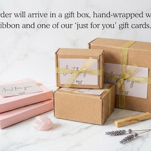 May include: Two light pink rectangular boxes and three brown cardboard boxes tied with gold ribbon. Each box features a white gift card with the text 'Just for you'. A raw rose quartz crystal, a wooden stick, and dried lavender sprigs are included.