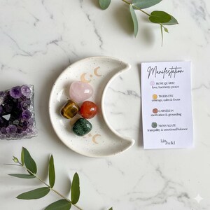 Peut inclure: Un plat en c&eacute;ramique blanc en forme de croissant de lune avec cinq pierres pr&eacute;cieuses polies : quartz rose, &oelig;il de tigre, cornaline et agate mousse. Une carte avec le mot "Manifestation" et des descriptions de pierres est &agrave; c&ocirc;t&eacute;.