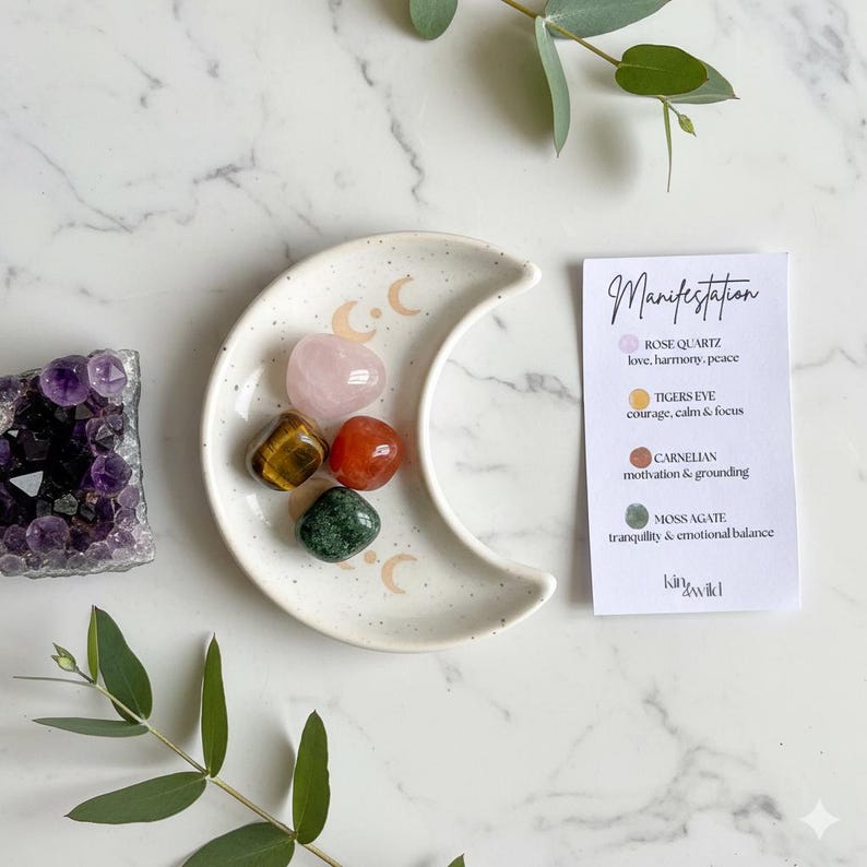 Peut inclure: Un plat en c&eacute;ramique blanc en forme de croissant de lune avec cinq pierres pr&eacute;cieuses polies : quartz rose, &oelig;il de tigre, cornaline et agate mousse. Une carte avec le mot "Manifestation" et des descriptions de pierres est &agrave; c&ocirc;t&eacute;.