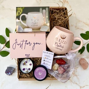 以下が含まれることがあります： 「Fueled by Crystals & Coffee」と書かれたピンク色のマグカップ、キャンドル、クリスタル、そして「Just for you...」と書かれたカードが入ったギフトボックス。ボックスには様々なアイテムが入っています。