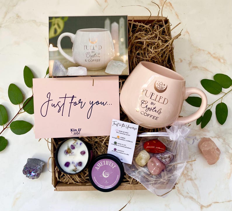 以下が含まれることがあります： 「Fueled by Crystals & Coffee」と書かれたピンク色のマグカップ、キャンドル、クリスタル、そして「Just for you...」と書かれたカードが入ったギフトボックス。ボックスには様々なアイテムが入っています。