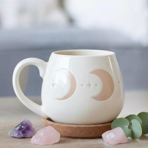 May include: Cream-coloured ceramic mug with a handle, decorated with crescent moons and stars. The mug rests on a cork coaster, with amethyst and rose quartz crystals. The mug has a rounded shape.