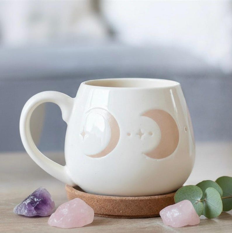 May include: Cream-coloured ceramic mug with a handle, decorated with crescent moons and stars. The mug rests on a cork coaster, with amethyst and rose quartz crystals. The mug has a rounded shape.