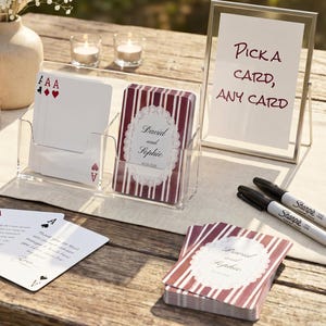 Op de afbeelding: Een doorzichtige acryl kaarthouder met speelkaarten met rood-witte strepen, gepersonaliseerd met de namen David en Sophie. Een bordje in een zilveren lijst toont de tekst Pick a card, any card, naast twee zwarte stiften en waxinelichtjes.