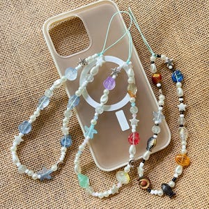 May include: Three beaded phone charms with a variety of colorful beads, including pearls, stars, and sea-themed shapes. The charms are attached to a clear phone case, resting on a textured, tan surface.
