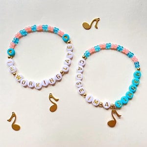 May include: Two beaded bracelets with pink and blue beads and white letter beads spelling out "I'm working late cause I'm a singer". Each bracelet has a gold musical note charm.