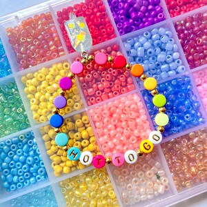 May include: A colorful beaded bracelet with the words "HOT TO GO" spelled out in white letters with gold outlines. The bracelet is made with a variety of colorful beads, including pink, blue, yellow, green, and red. The bracelet is surrounded by small, colorful beads in a clear plastic container.