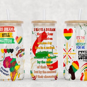 May include: Three glass tumblers with bamboo lids and straws. Each tumbler features a different design with text and graphics celebrating Black History. The designs include phrases like "His Dream Still Matters" and "I Have a Dream".