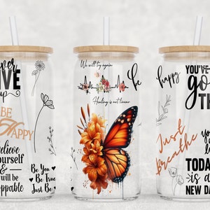 May include: Three glass tumblers with bamboo lids and straws. Each tumbler features a different inspirational quote and design. The first tumbler has a black and white design with the quote "never give up, be happy, believe in yourself & you will be unstoppable". The second tumbler has a butterfly and floral design with the quote "We will try again, healing is not linear". The third tumbler has a black and white design with the quote "You've got this, just breathe, you are loved, today is a new day".