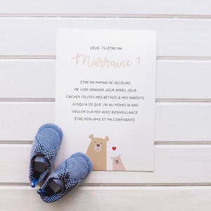May include: A white card with the text 'VEUX-TU ETRE MA Marraine?' in pink script. The text below reads 'ÊTRE MA MAMAN DE SECOURS ME VOIR GRANDIR JOUR APRÈS JOUR CACHER TOUTES MES BETISES À MES PARENTS JUSQU'À CE QUE J'AI AU MOINS 18 ANS VEILLER SUR MOI AVEC BIENVEILLANCE ÊTRE MON AMIE ET MA CONFIDENTE'. The card is decorated with two brown bears and a pink heart. Two blue baby shoes with white bows are on a white wooden surface.