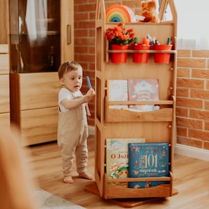 May include: A wooden toy shelf with a house-shaped design. The shelf has multiple shelves for books and toys, as well as a small area for potted plants. A young child is standing next to the shelf, holding a blue marker.