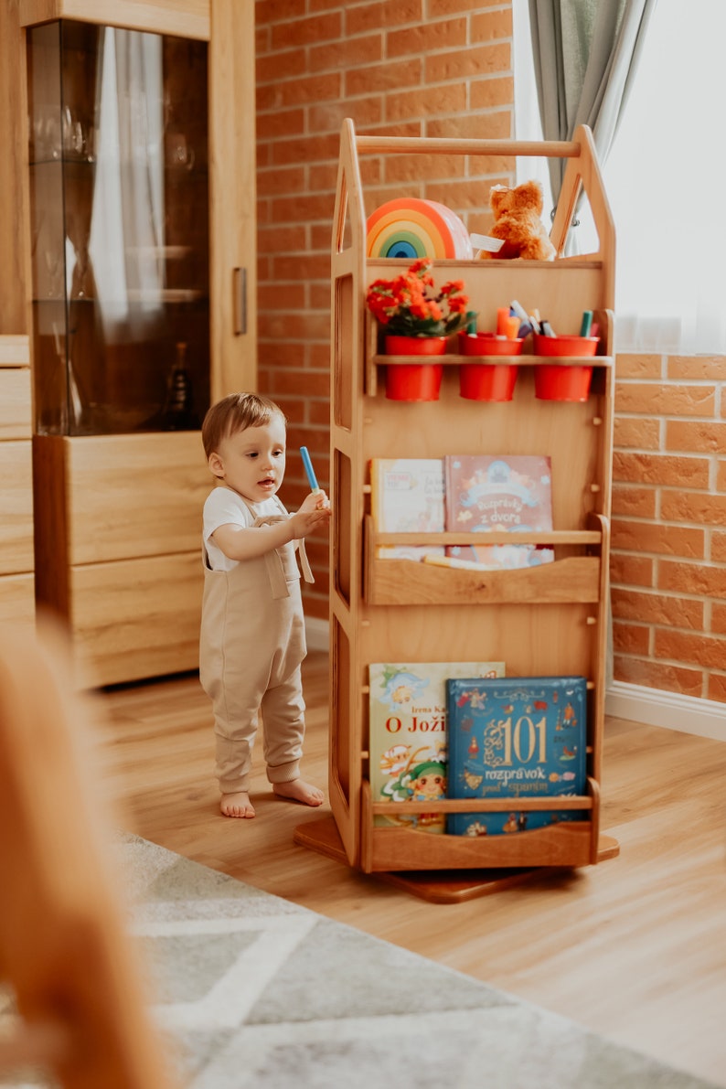 May include: A wooden toy shelf with a house-shaped design. The shelf has multiple shelves for books and toys, as well as a small area for potted plants. A young child is standing next to the shelf, holding a blue marker.