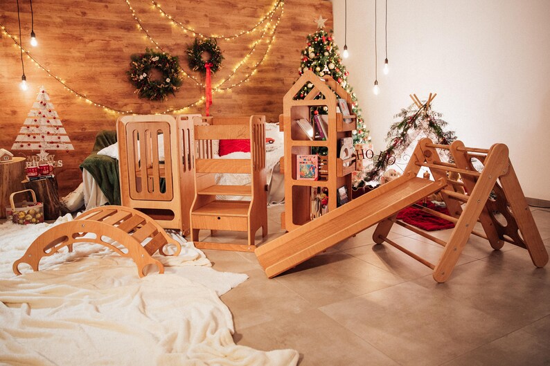 May include: A set of wooden children's toys, including a slide, a climbing frame, a rocking horse, and a bookshelf. The toys are arranged in a room with a wooden floor and a white rug. There are Christmas decorations in the background.