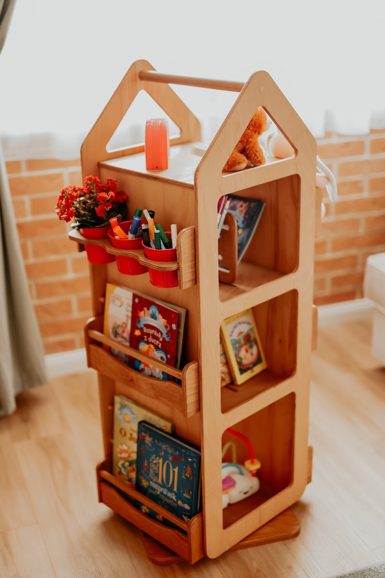 May include: A wooden toy bookshelf shaped like a house with a red roof. The bookshelf has four shelves and a small drawer. The shelves are filled with books and toys. There is a small potted plant on the top shelf.