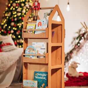 May include: A wooden bookshelf shaped like a house with a rotating base. The bookshelf has four shelves and is filled with colorful children's books. A nutcracker doll is on top of the bookshelf.