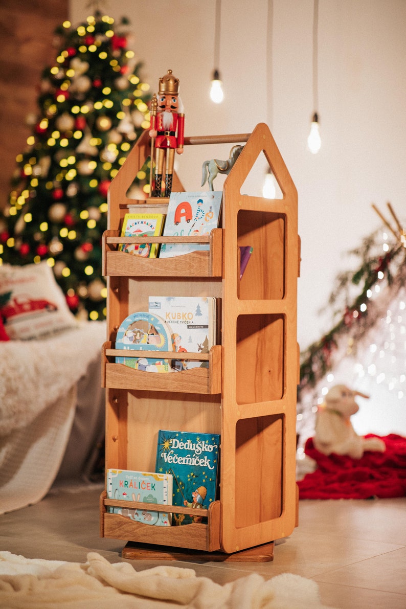 May include: A wooden bookshelf shaped like a house with a rotating base. The bookshelf has four shelves and is filled with colorful children's books. A nutcracker doll is on top of the bookshelf.