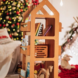 May include: A wooden bookshelf shaped like a house with a nutcracker figurine on top. The bookshelf has four shelves filled with children's books and a rocking horse toy. The bookshelf is in front of a Christmas tree.