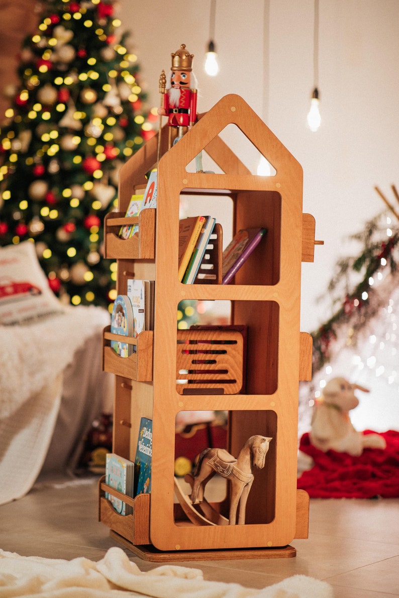 May include: A wooden bookshelf shaped like a house with a nutcracker figurine on top. The bookshelf has four shelves filled with children's books and a rocking horse toy. The bookshelf is in front of a Christmas tree.