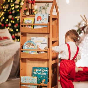 May include: A wooden bookshelf with a house-shaped design, featuring four shelves filled with colorful children's books. A nutcracker figurine and a wooden horse are on top of the bookshelf. A young child is standing next to the bookshelf, reaching for a book.