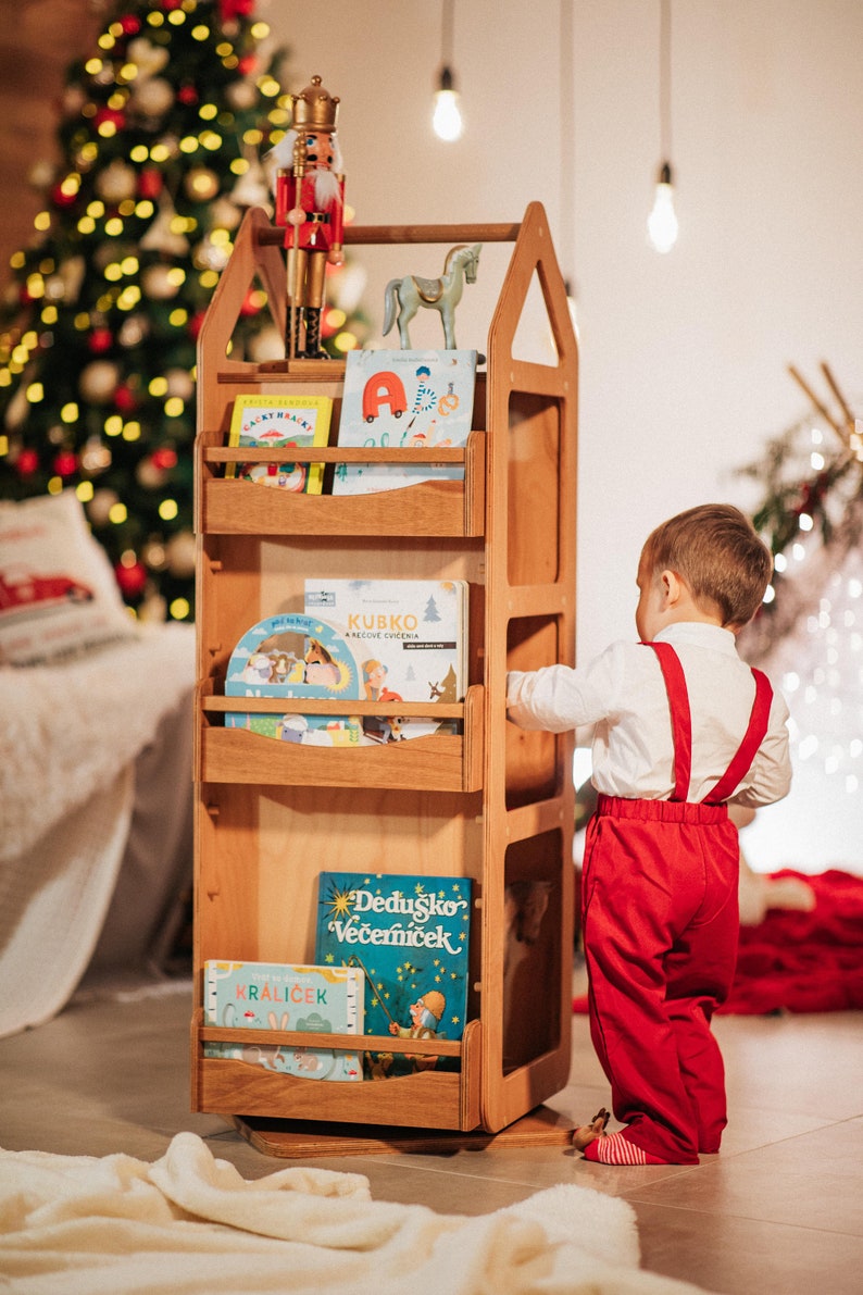 May include: A wooden bookshelf with a house-shaped design, featuring four shelves filled with colorful children's books. A nutcracker figurine and a wooden horse are on top of the bookshelf. A young child is standing next to the bookshelf, reaching for a book.