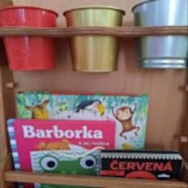 May include: A wooden shelf with three metal buckets in red, gold, and silver. Below the buckets are three books, one with the title "Barborka" in pink letters, another with a monkey illustration, and a third with the word "CERVENA" in red letters.