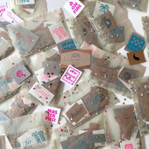 Könnte beinhalten: Eine Sammlung kleiner, bunter und gemusterter Bügelbilder in durchsichtigen Plastiktüten. Die Tüten sind mit goldenem Glitzer gefüllt. Die Bügelbilder zeigen verschiedene Designs, darunter ein Peace-Zeichen, eine Palme, ein kariertes Muster, einen Schnurrbart, ein Herz und die Worte "Sew Much Love".