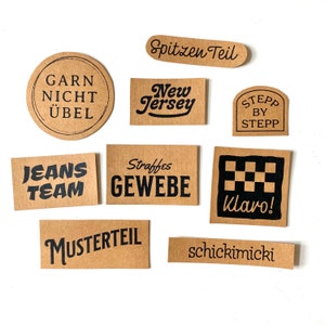 Könnte beinhalten: Neun braune Papieretiketten mit schwarzem Text. Die Etiketten zeigen Sätze wie "Garn Nicht Uebel", "New Jersey", "Straffes Gewebe", "Jeans Team", "Musterteil", "Spitzen Teil", "Stepp by Stepp", "Klaro!" und "schickimicki".