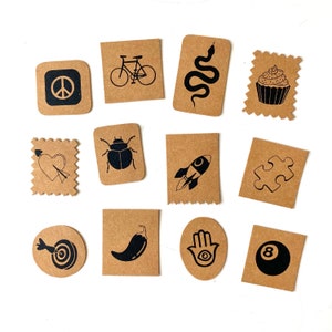 Könnte beinhalten: Satz von 10 braunen Papierkarten mit schwarzen Illustrationen. Die Illustrationen beinhalten ein Peace-Zeichen, ein Fahrrad, eine Schlange, einen Cupcake, ein Herz mit einem Pfeil, einen Käfer, eine Rakete, ein Puzzle-Teil, eine Chilischote, eine Hamsa-Hand und eine Billardkugel.