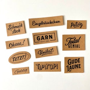 Könnte beinhalten: Eine Ansammlung brauner rechteckiger und ovaler Etiketten mit schwarzer Schrift. Die Etiketten zeigen verschiedene deutsche Wörter und Sätze, darunter "Schmuckstück", "Einzelstückchen", "Ohaaa!", "GARN", "Unikat", "FETZT!", "Goldig", "TIPITOP", "Gude Laune", "Putzig", und "Total Genial".
