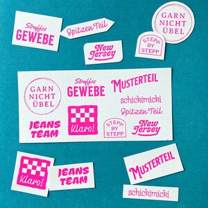 Könnte beinhalten: Eine Sammlung von rosa Gummistempeln mit verschiedenen Designs und Texten, darunter "Straffes Gewebe", "Spitzen Teil", "New Jersey", "Garn Nicht Uebel", "Jeans Team", "Klaro!" und "Musterteil".